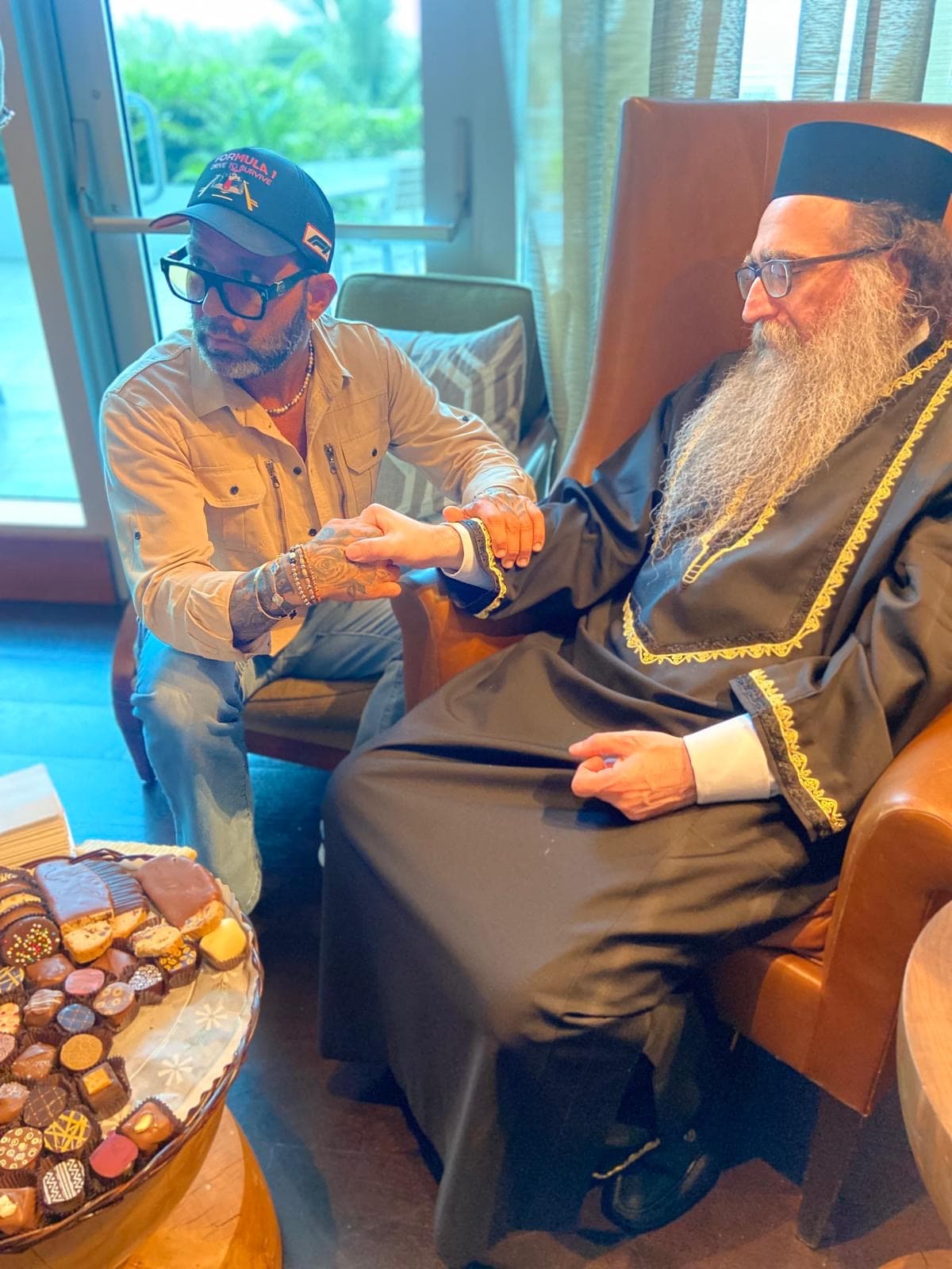 Tattooed man in a cap holds the hand of a bearded religious leader in robes.