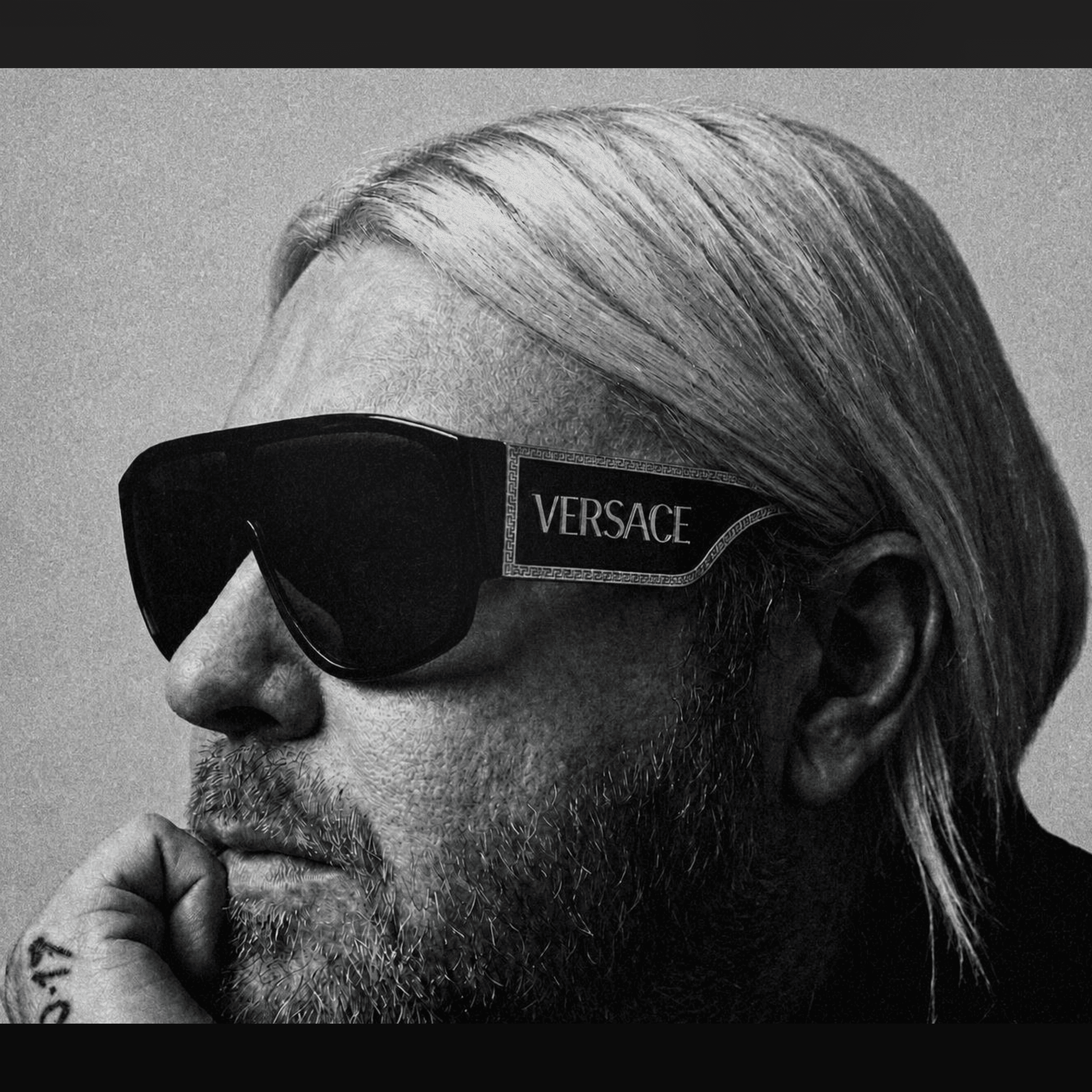 Monochrome profile of a bearded man with long hair wearing oversized black Versace sunglasses.